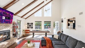 A modern living room with a fireplace, a large gray sofa, a coffee table, and a colorful rug. There is a bookshelf with books and decorative items, and a large window with a view of the outside. - The Ultimate Getaway: Pool, Pickleball, Mini Golf (San Antonio)