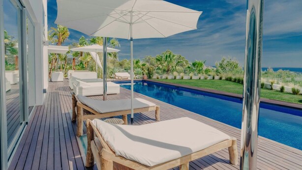 Terraza de madera con sillones de descanso y sombrillas, con vistas a una piscina.