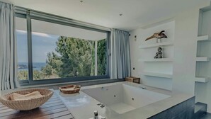 Un baño moderno con una bañera grande, una mesita de madera y vistas al mar a través de un amplio ventanal.