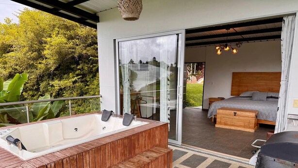 A modern outdoor bathroom with a wooden bench, a hot tub, and a glass door leading to a bedroom with a bed and a wooden headboard. - Alto del sion  peñol guatape  (El Peñol)