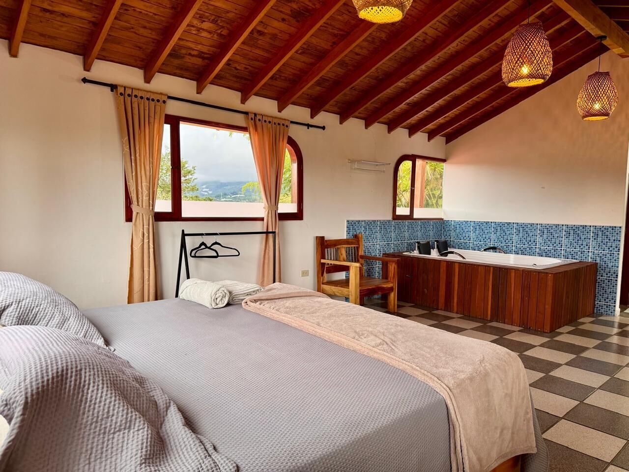 Un dormitorio con dos camas, techo de madera, ventana con cortinas, una silla, un escritorio pequeño y un baño con lavamanos y pared de azulejos azules.