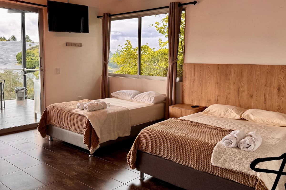 A hotel room with two beds, a TV mounted on the wall, a balcony with a view of greenery, and a wooden headboard.