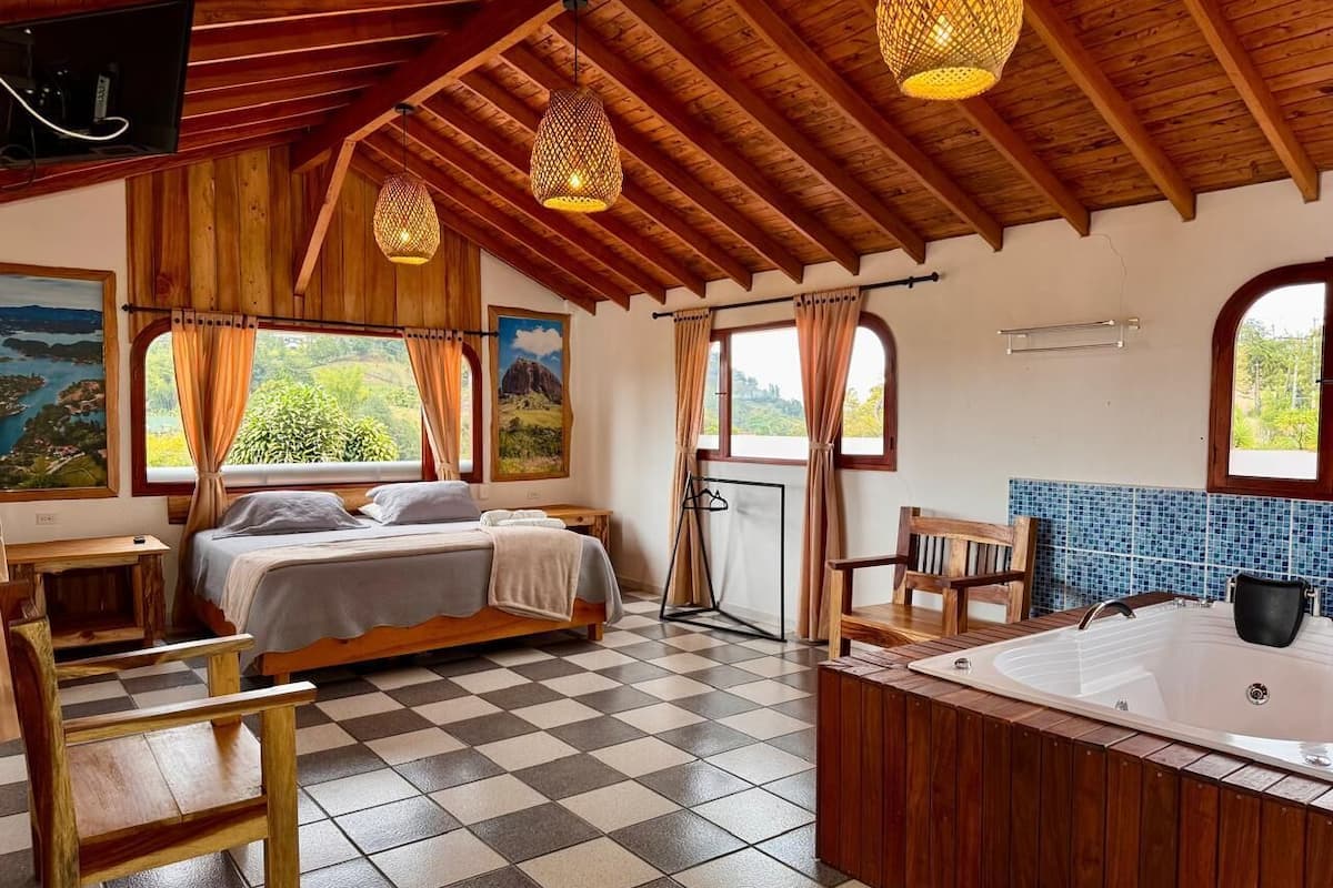 A room with a bed, a wooden bench, a bathtub, and a view of greenery through the windows.