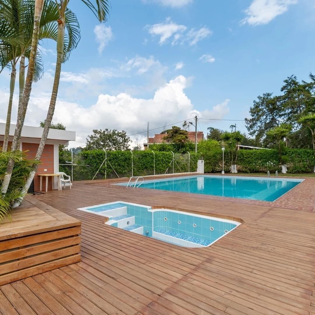 A rectangular swimming pool with a wooden deck surrounding it, a small seating area, and a building in the background.