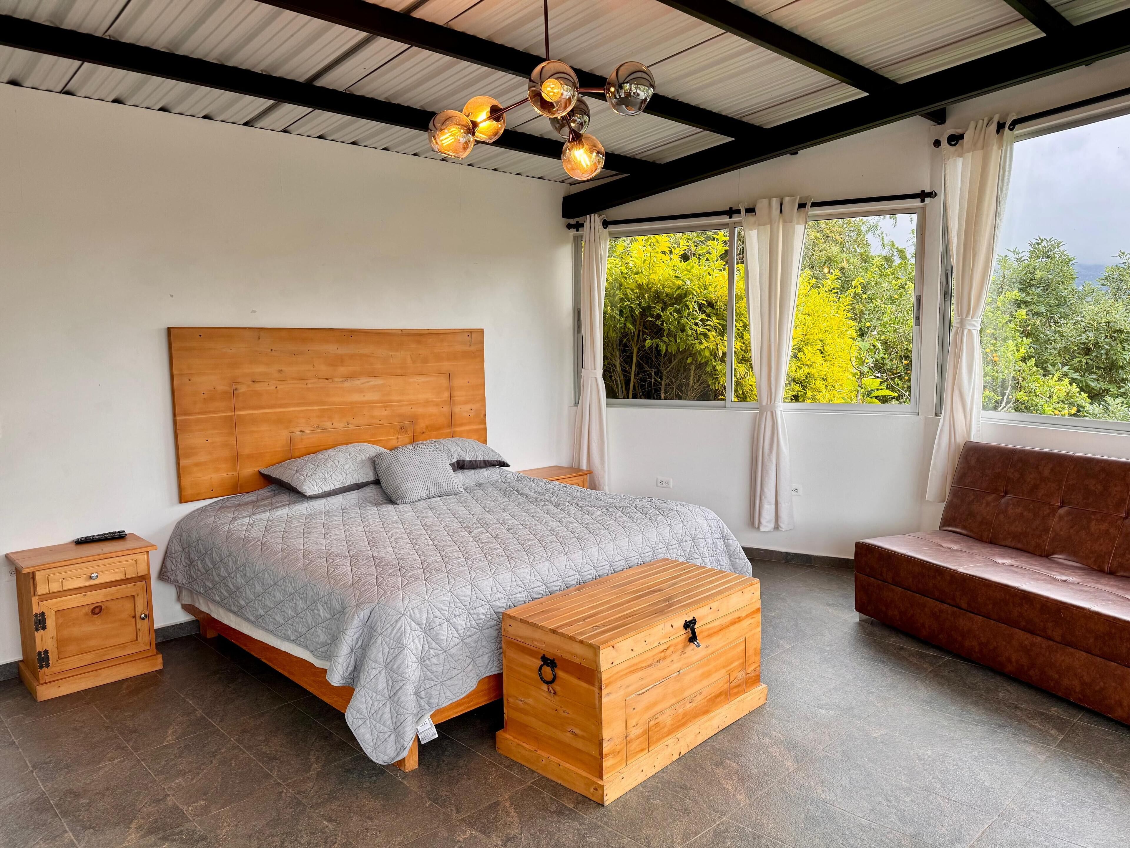A bedroom with a wooden headboard, a bed with a grey quilt, a wooden chest, a bench, and a window with curtains.