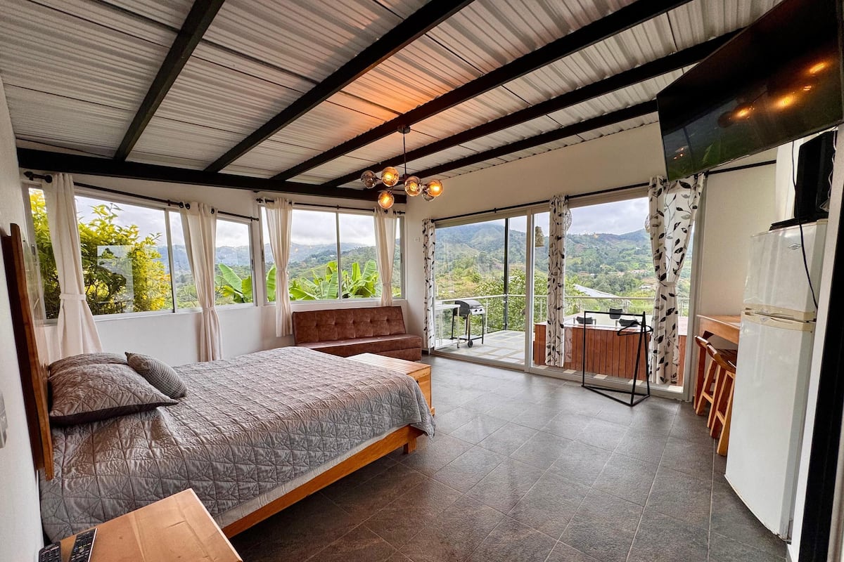 A bedroom with a large bed, a wooden nightstand, a TV, and a balcony with a view of the mountains.