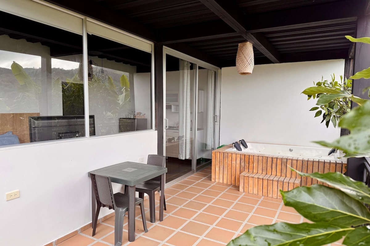 A patio area with a table and chairs, a wooden hot tub, and a large window overlooking a natural landscape.