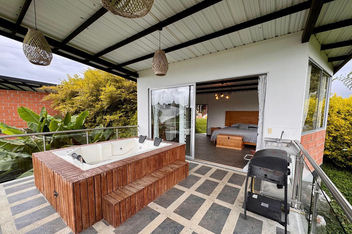 A modern outdoor bathroom with a wooden tub, a barbecue grill, and a glass-enclosed bedroom with a bed and a nightstand.