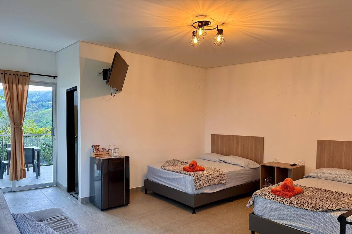 A hotel room with two beds, a sofa, a TV mounted on the wall, and a balcony with a view of the hills.