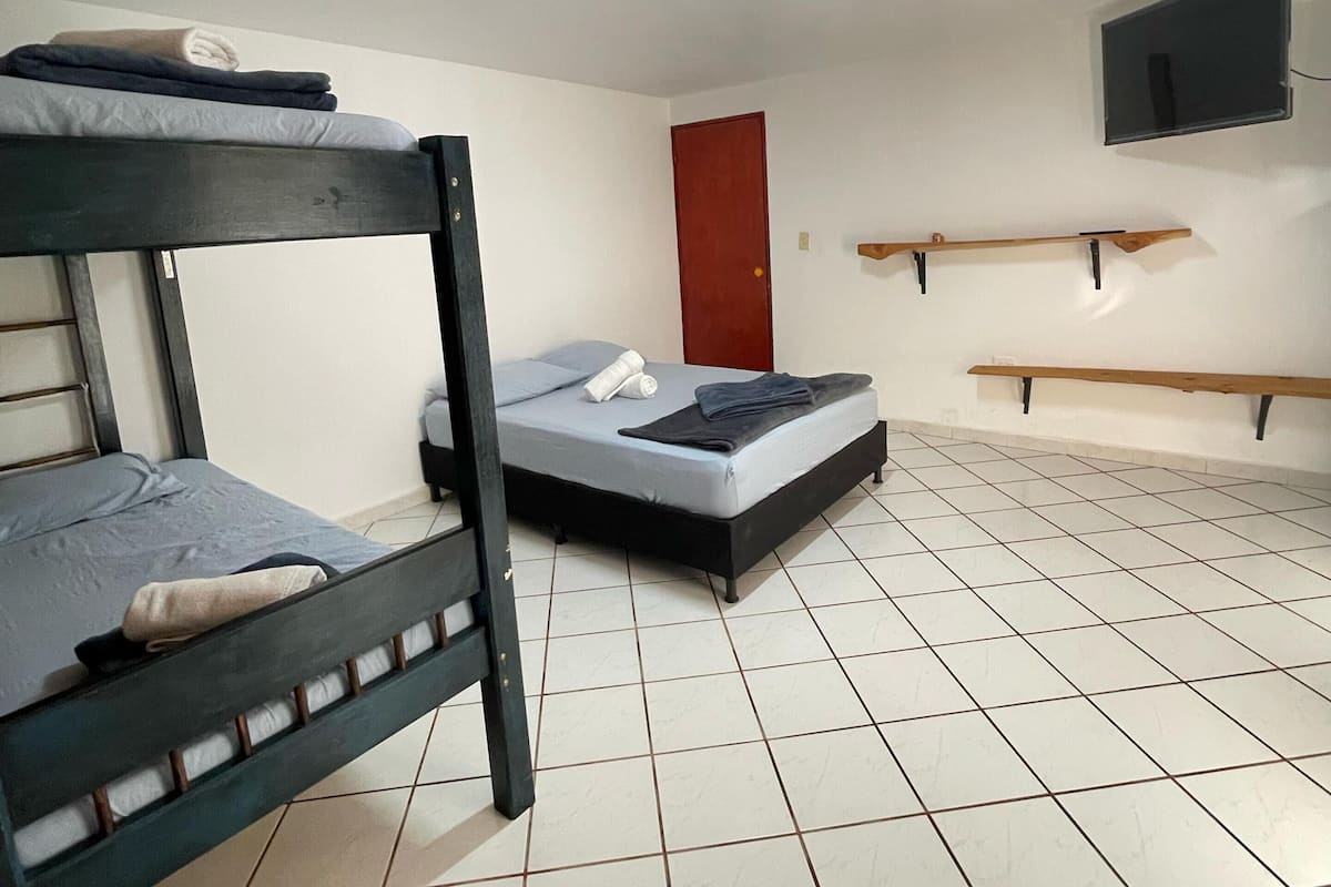 A dormitory room with two bunk beds, a single bed, tiled floor, a red door, a mounted TV, and a wooden shelf.