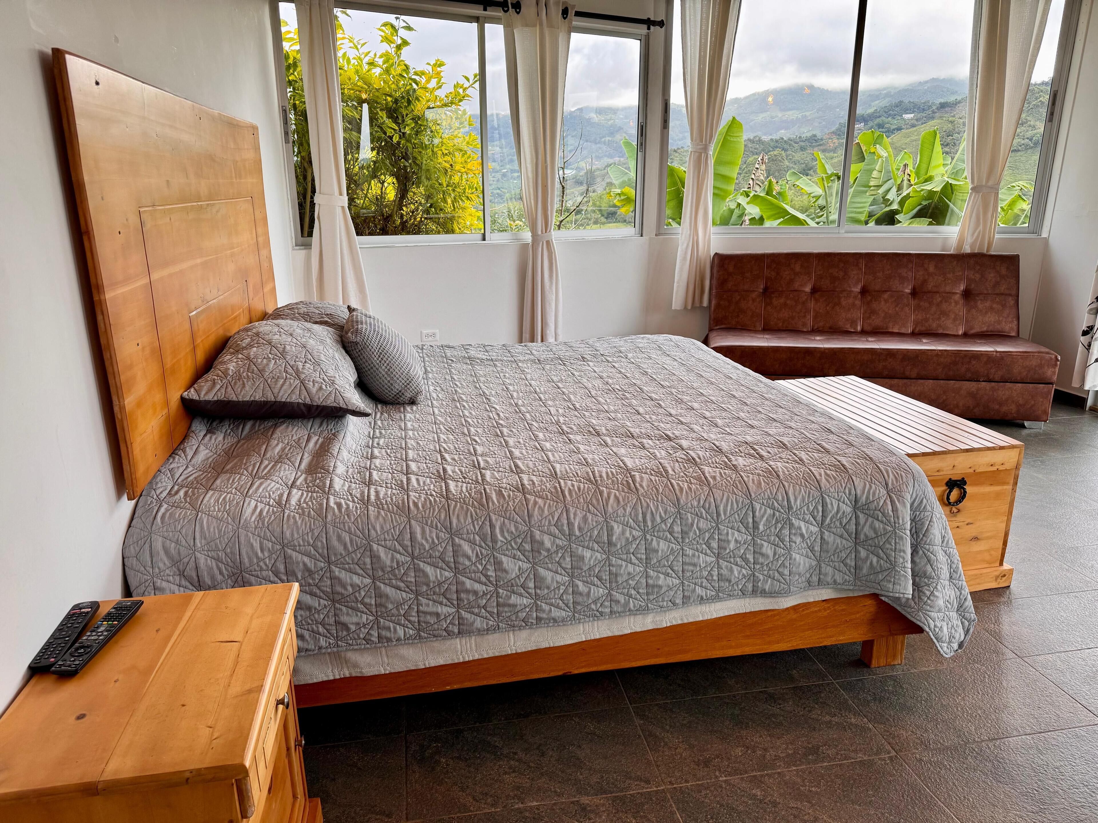 A bedroom with a wooden bed, a wooden nightstand, a brown sofa, and a large window with a view of greenery and mountains.