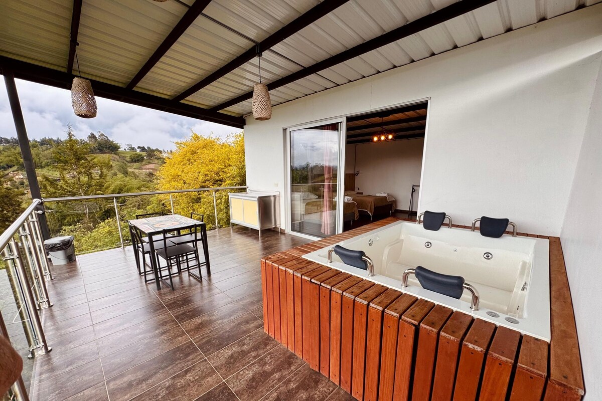 A balcony with a wooden table and chairs, a hot tub, and a glass door leading to a bedroom.