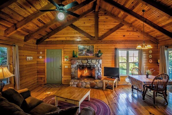 A cozy log cabin interior with a stone fireplace, wooden furniture, and a television.