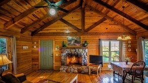 A log cabin interior with a stone fireplace, wooden furniture, and a ceiling fan.