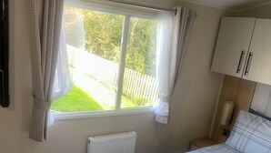 A bedroom with a bed, a window with curtains, a radiator, and a wooden nightstand with a lamp. - Charming 2-bed Chalet in Tuxford (Newark)