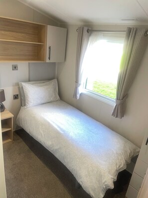 A single bed with a white bedspread, a wooden shelf, a bedside table, a window with curtains, and a small wardrobe. - Charming 2-bed Chalet in Tuxford (Newark)