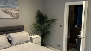 Een moderne slaapkamer met een bed, een nachtkastje, een kamerplant en een badkamer zichtbaar door een geopende deur.