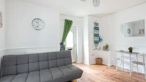 A modern living room with a grey sofa, a dining area with a small table and chairs, a clock on the wall, and a green curtain. - Cheerful Maisonette in the Heart of Orrell Park (Liverpool)
