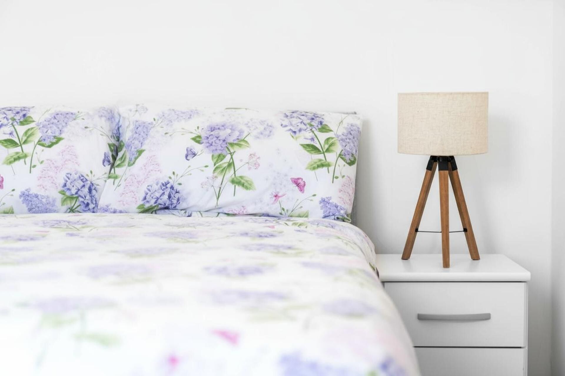 Una cama bien hecha con un edredón estampado con motivos florales, una mesita de noche blanca con una lámpara y un cajón.