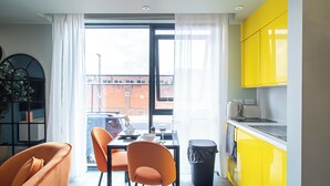 A modern kitchen with yellow cabinets, a dining area with orange chairs, and a view of a brick building through large windows. - Sheffield City Escape - Modern Apartment (Sheffield)