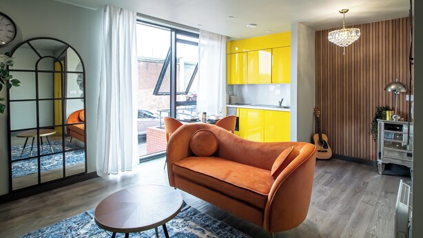 A modern living room with a sofa, a small round table, and a view of a bright yellow kitchen through a glass door. - Sheffield City Escape - Modern Apartment (Sheffield)