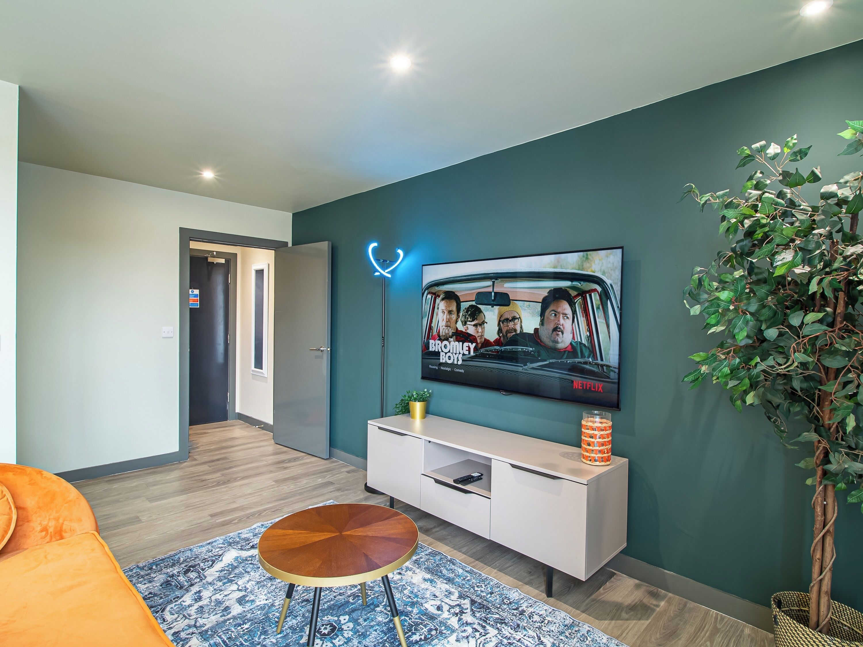 Una sala de estar moderna con un televisor de pantalla plana montado en la pared, un mueble blanco debajo, una mesa de centro redonda de madera, un tapete estampado y una planta en maceta.