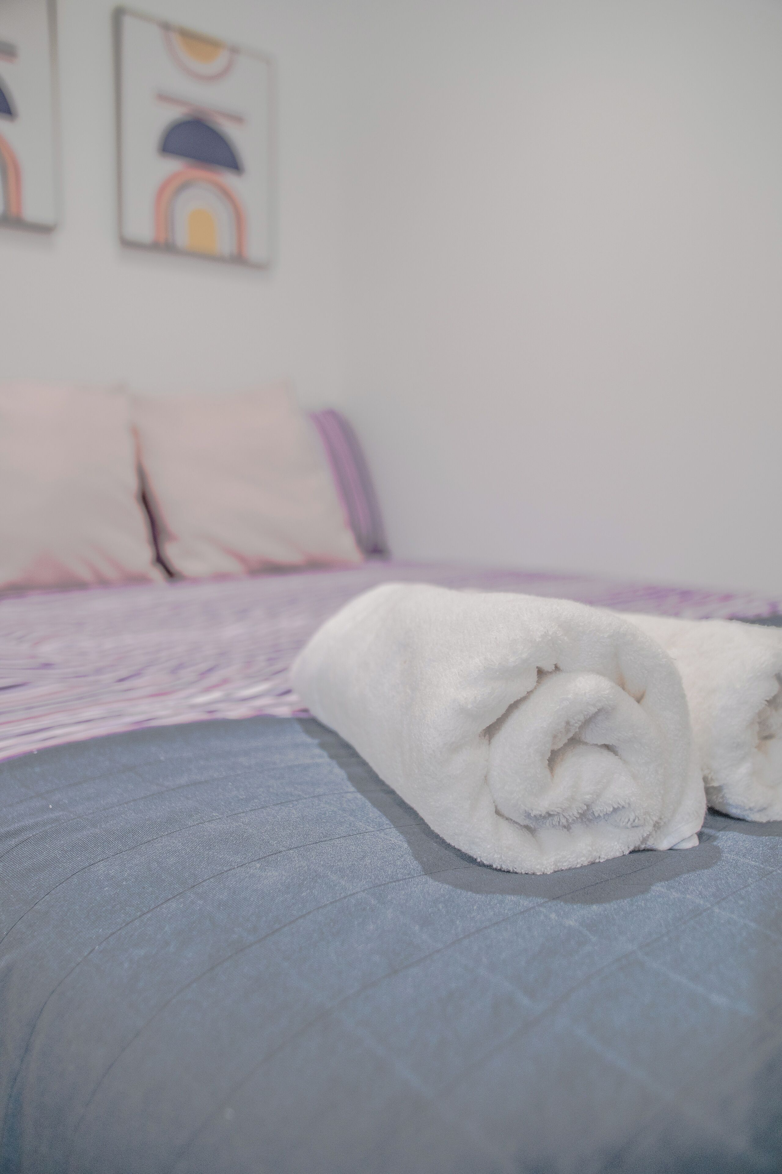 Ein ordentlich bezogenes Bett mit einem aufgerollten weißen Handtuch, einer violetten Decke und einer Wand mit abstrakter Kunst.