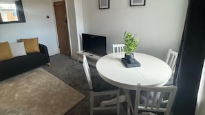 Una sala de estar pequeña y moderna con una mesa redonda blanca, dos sillas blancas, un sofá negro con cojines mostaza y blancos, un televisor sobre un mueble bajo y una planta en maceta sobre la mesa.