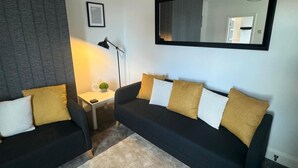 Un moderno salón con un sofá negro, una pequeña mesita auxiliar blanca con una lámpara y un espejo en la pared.