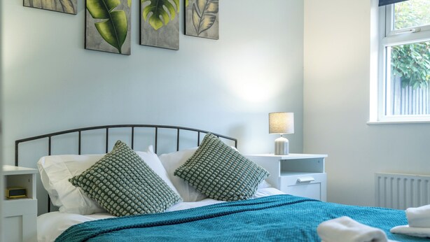 A neatly made bed with a teal blanket and two patterned pillows, a bedside lamp, and artwork on the wall. - Crawley Stainer 1-bedroom With Parking and Garden (Crawley)