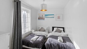 A twin bedroom with two beds, a window with curtains, a hanging light fixture, and two wall art pieces. - 3 Bed Home w/ 2 Bathrooms &free Parking Sleeps 6 (London)
