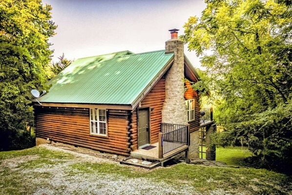 A log cabin with a green metal roof, a stone chimney, and a small porch with a black railing.