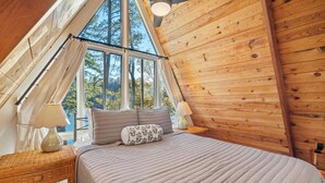 Un dormitorio acogedor con un ventanal grande, paredes de madera, una cama con una colcha a rayas y dos mesitas de noche con lámparas.