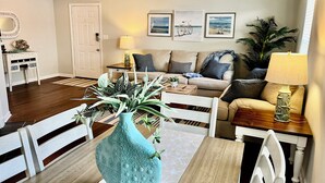 Ein Wohnzimmer mit Esstisch, Stühlen, einer Couch, einem Couchtisch mit Pflanzen, einer Lampe und gerahmten Bildern an der Wand.