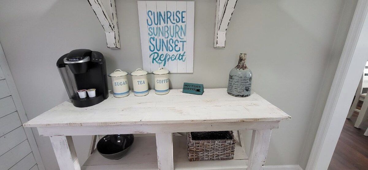 A coffee station with a Keurig machine, sugar, tea, and coffee containers, a bottle, and a sign that reads 'SUNRISE SUNBURN SUNSET Repeat.'