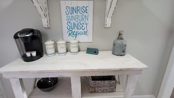 A coffee station with a Keurig machine, sugar, tea, and coffee containers, a bottle, and a sign that reads 'SUNRISE SUNBURN SUNSET Repeat.'