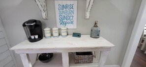 Un coin café équipé d’une machine Keurig, de sucre, de sachets de thé et de dosettes de café, d’une bouteille d’eau et d’une pancarte sur laquelle est écrit : « LEVER DU SOLEIL, COUP DE SOLEIL, COUCHER DU SOLEIL. Répéter. »
