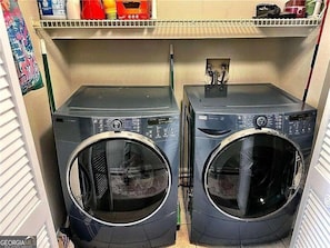 Two front-loading washing machines in a laundry room with a shelf above containing various items.