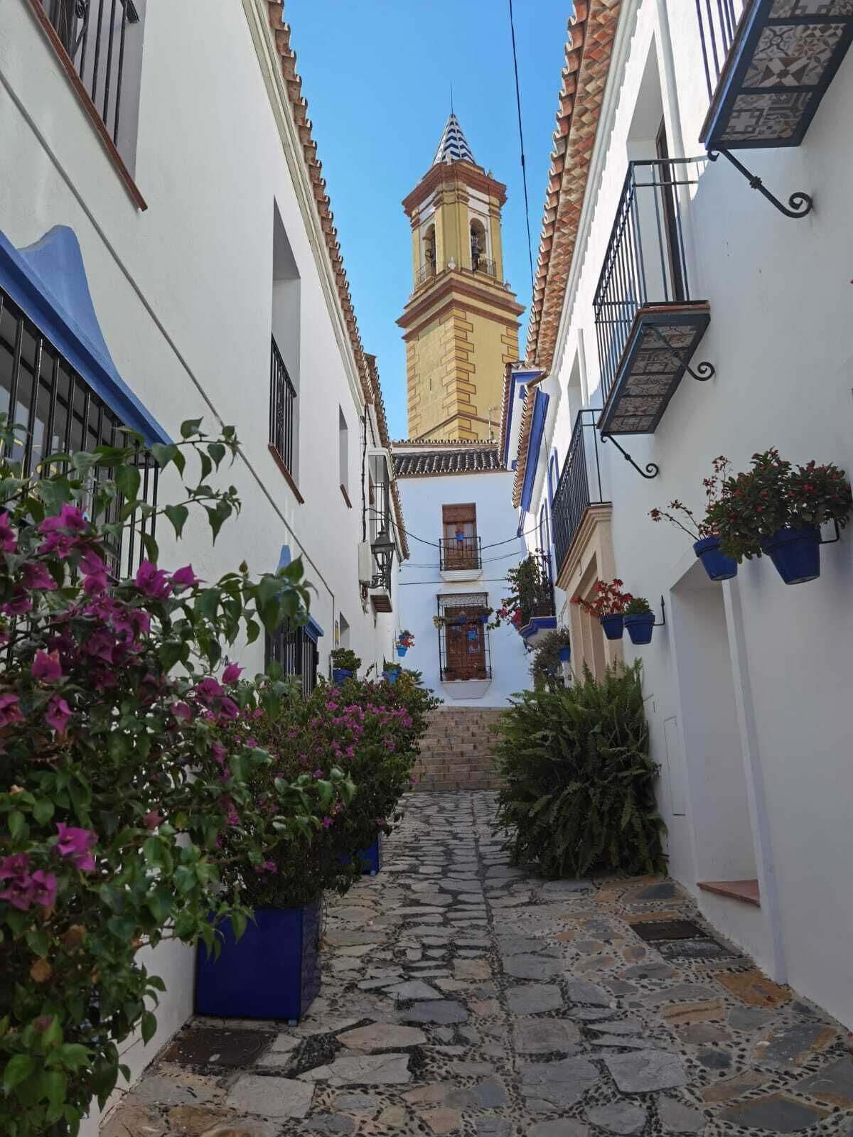 Una estrecha calle adoquinada flanqueada por edificios blancos con toldos azules y balcones, que conduce a una torre con una campana.