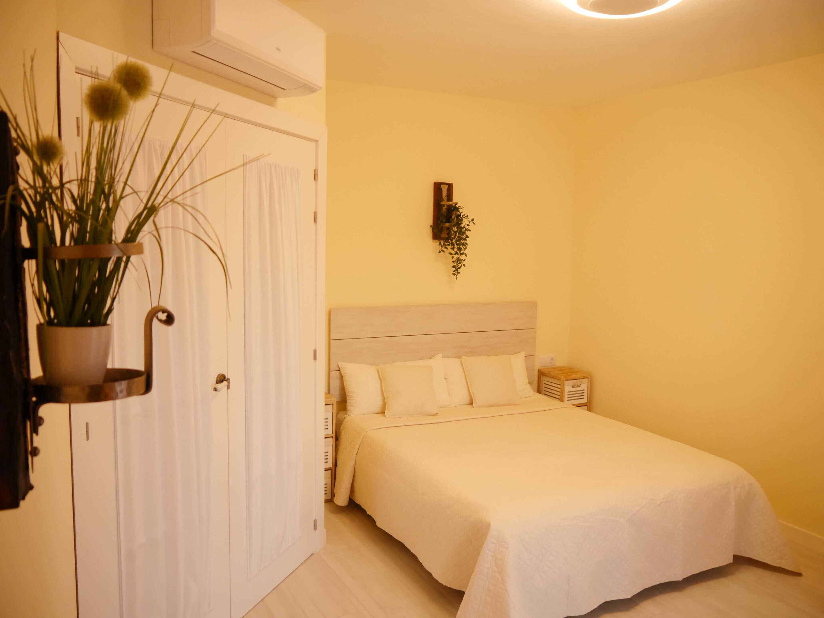 A bedroom with a bed, a potted plant, a nightstand, and an air conditioning unit.