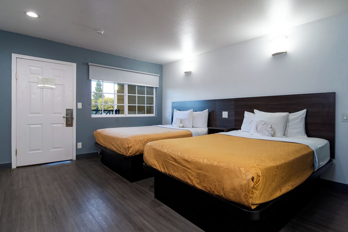A hotel room with two beds, a window with blinds, a door, and a light fixture on the ceiling.