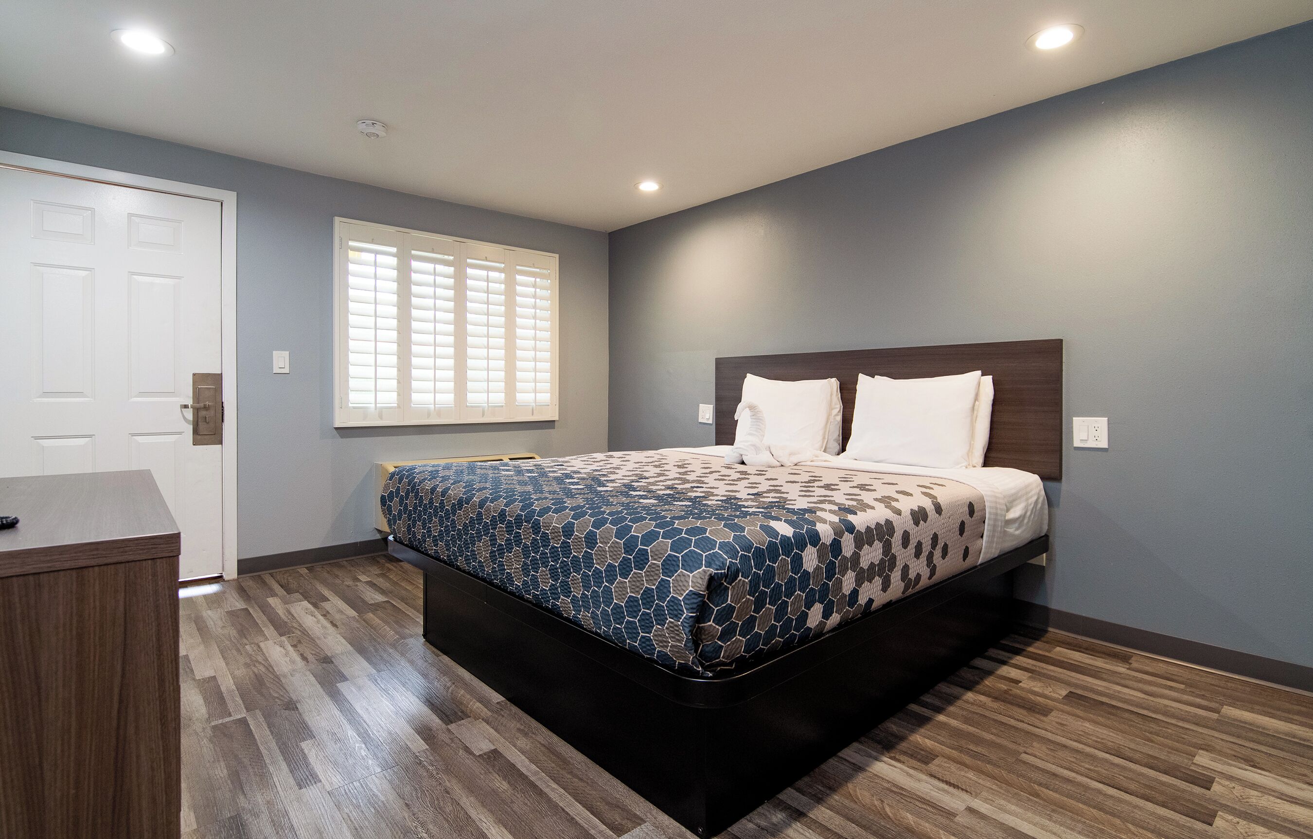 A bedroom with a bed, a desk, a window with blinds, and a door.