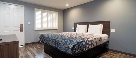 A bedroom with a bed, a desk, a window with blinds, and a door.