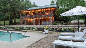 A pool area with lounge chairs and a building with outdoor lighting. - Inground Pool, Hot Tub, Fire Pit at Large Log Cabin near Mohican State Park (Butler)
