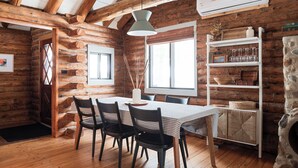 A rustic wooden cabin interior with a dining table, chairs, a white shelf with glassware, and a stone fireplace. - Inground Pool, Hot Tub, Fire Pit at Large Log Cabin near Mohican State Park (Butler)