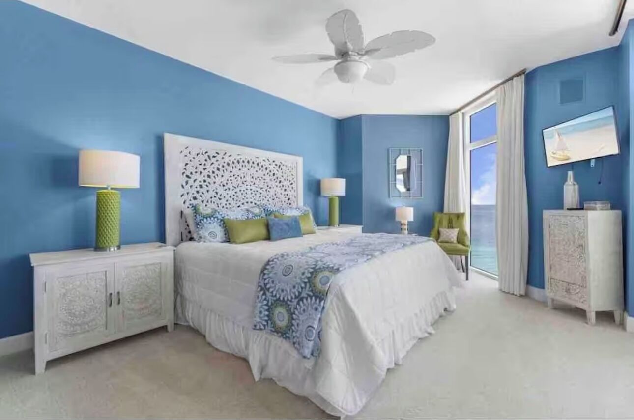 A bedroom with a bed, nightstands, a ceiling fan, a window with curtains, and a television.