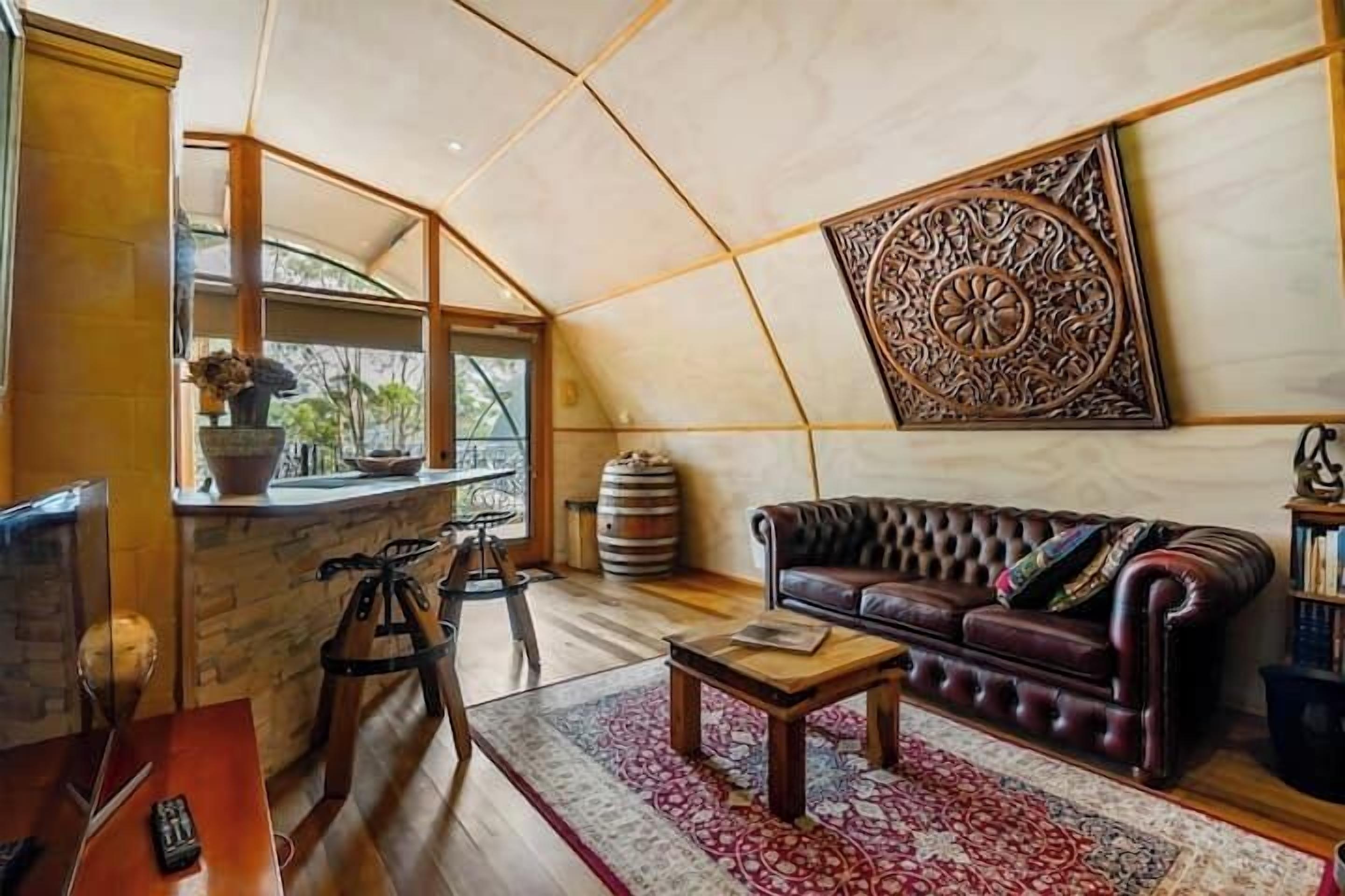A cozy living area with a leather sofa, a coffee table, and a patterned rug. There is a large decorative wall piece and a window with a view of greenery.