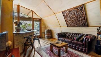 A cozy living area with a leather sofa, a coffee table, and a patterned rug. There is a large decorative wall piece and a window with a view of greenery.
