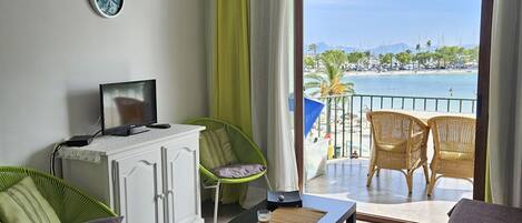 A room with a ceiling fan, a TV on a cabinet, a sofa, a table with a glass, and a view of a beach and boats through a large window.