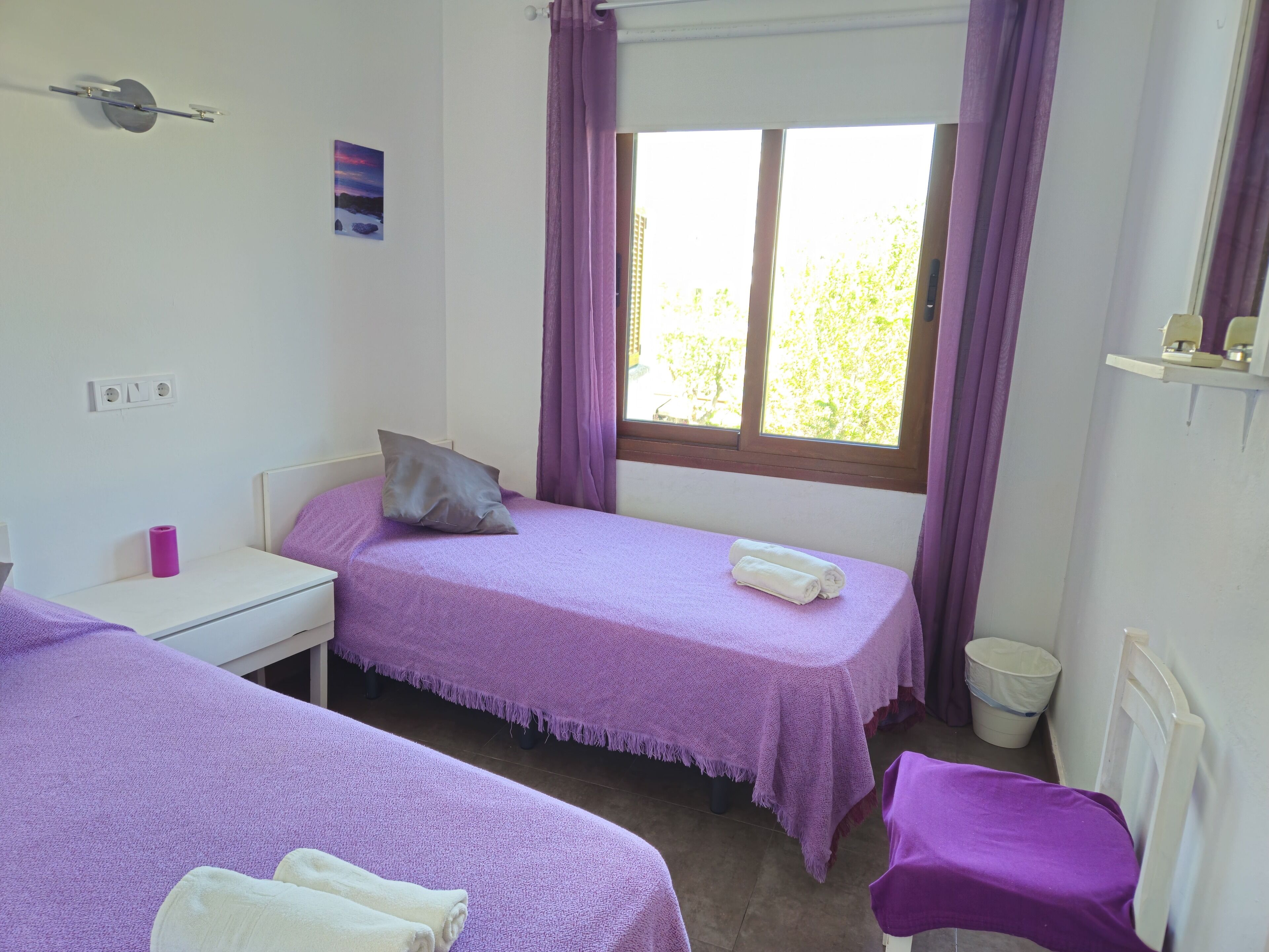 A small, clean room with two beds, a window with curtains, a desk, and a small trash bin.
